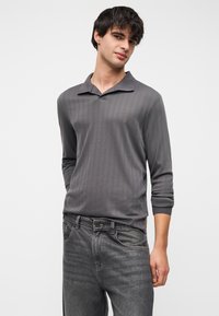 Polo gris à manches longues avec col, présentant une texture côtelée verticale. Associé à un jean en denim gris foncé. Design simple sans motifs supplémentaires.