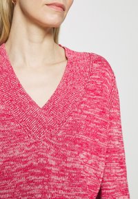 Roze gebreide trui met V-hals en een textuurpatroon, met een losse pasvorm en geribbelde afwerking. Zacht stof met een gemêleerd kleureffect.