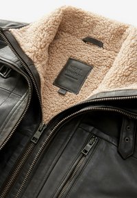 Black leather jacket with beige sherpa lining, front zipper partially open, zippered chest pocket, and "Next Signature" label inside collar.