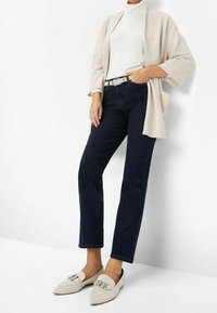 Chaqueta kimono beige sobre un suéter blanco de cuello alto, combinada con jeans de denim oscuro y mocasines gris claro con detalles de hardware plateado.