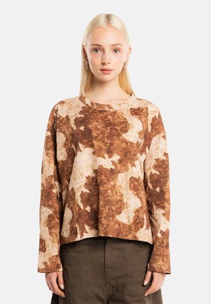 Joven mujer con pelo rubio liso lleva una camisa de manga larga con estampado de vaca en tonos marrón y beige y pantalones oscuros, de pie frente a un fondo sencillo.