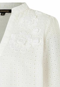 Weiße Bluse mit einem strukturierten Muster, das kleine Quadrate und florale Stickereien auf der Brust aufweist. Hoher Kragen.