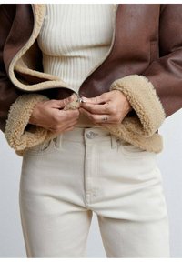 Chaqueta marrón con cremallera y forro de pelaje beige, usada sobre una blusa ribeteada crema y unos pantalones vaqueros beige claros, con las manos agarrando la cremallera.