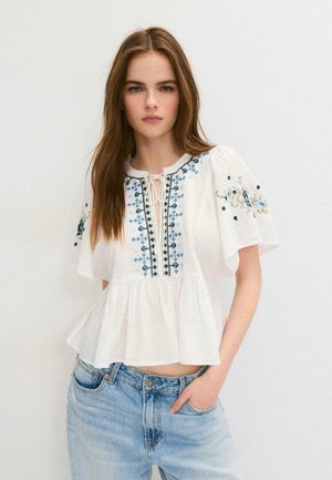 Jeune femme aux longs cheveux bruns portant un chemisier blanc brodé à manches courtes et un jean bleu clair, posant devant un fond uni.