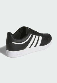 Zapatilla deportiva negra con diseño de tres rayas blancas, parte superior de cuero y textil suave, cuello acolchado y suela de goma blanca.