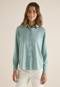 Femme souriante portant une chemise à manches longues bleu clair boutonnée et un pantalon blanc cassé, debout devant un fond neutre.