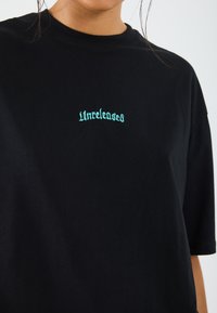 Black cotton t-shirt with short sleeves featuring turquoise embroidered text "Unreleased" on the chest. Simple and relaxed fit.