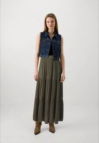 Gilet in denim con un lavaggio blu scuro e tasche frontali, abbinato a una lunga gonna tiered verde oliva e stivaletti marroni.
