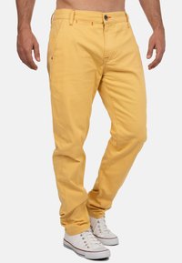 Chino gialli in cotone con passanti per cintura, chiusura a bottone e cuciture arancioni in contrasto, abbinati a sneakers bianche.