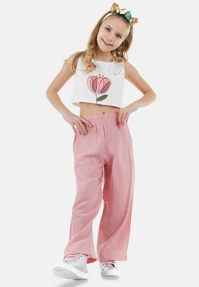 Witte crop top met een roze bloemenafdruk, gecombineerd met lichtroze, wijde, gestructureerde broek. Model draagt witte sneakers en een bloemenhoofdband.
