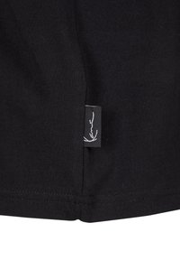 Black cotton T-shirt hem with a white label featuring a signature logo sewn into the side, smooth texture, and clean stitching.