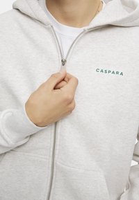 Lys grå hettegenser med glidelås laget av myk stoff. Har en frontglidelås, to lommer og grønn "CASPARA" logo på venstre brystområde.