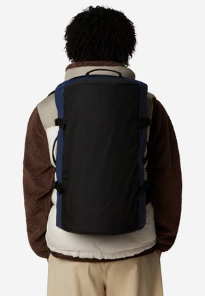 Zaino nero con accenti blu, caratterizzato da una forma rettangolare, spallacci regolabili e una maniglia superiore. Indossato sopra una giacca marrone e crema.