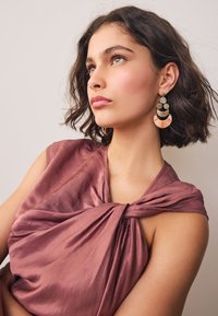 Gold and peach earrings feature circular shapes with intricate patterns and a textured finish. The model wears a mauve satin top with a knot detail.