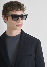 Crne sunčane naočale s angularnim, velikim okvirima. Model nosi tamni sako od teksturiranog materijala i tamnu majicu s okruglim izrezom ispod.