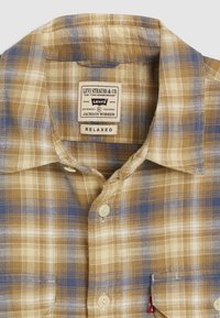 Camisa xadrez nas cores bege, azul e creme. Feita de um tecido macio, com colarinho, fecho de botão e uma pequena etiqueta com o logótipo no bolso.