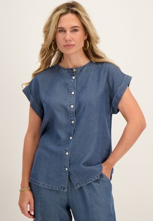 Vrouw met lang blond haar draagt gouden oorringen, een spijkerblouse met korte mouwen en knopen, en een bijpassende spijkerbroek, één hand in de zak.