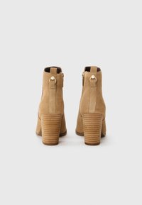 Bottines en suède tan avec un bout légèrement pointu, des accents de fermeture éclair dorée, et un talon en bois strié. Texture lisse et forme structurée.