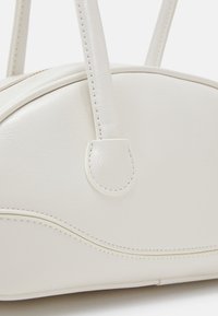 Gros plan d'un sac à main en cuir blanc à forme arrondie, coutures visibles et deux poignées robustes fixées sur le dessus.