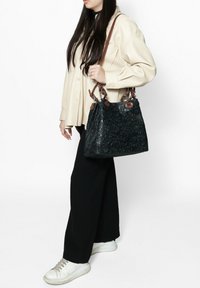 Borsa a tracolla nera in pelle con motivo floreale, manici in pelle marrone e forma strutturata, indossata con un top crema e pantaloni neri.