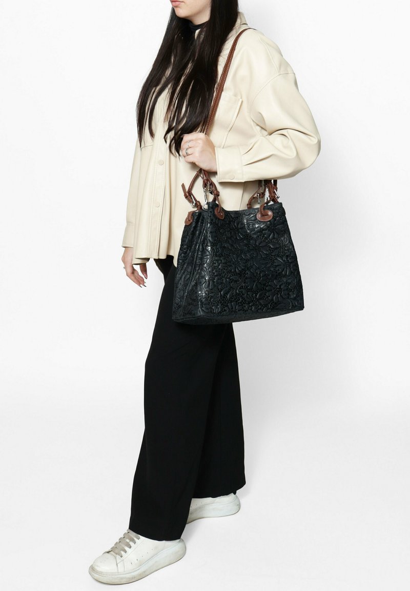 Borsa a tracolla nera in pelle con motivo floreale, manici in pelle marrone e forma strutturata, indossata con un top crema e pantaloni neri.