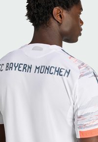 Biała koszulka FC Bayern München z siateczkową teksturą, kolorowymi akcentami na ramionach oraz wyrazistym napisem na plecach.