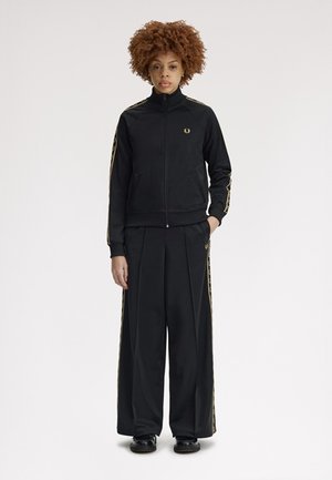 Jeune personne debout portant une veste de survêtement noire zippée et un pantalon à jambes larges avec des bandes dorées, mains détendues, expression neutre.