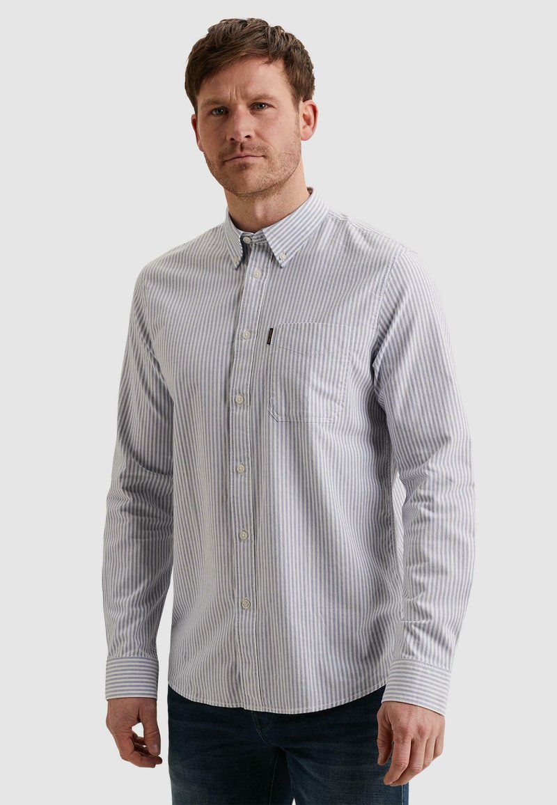 Muž nosiaci dlhorukú, zapínaciu svetlomodro-bielu pruhovanú košeľu s predným vreckom, stojaci pred jednoduchým pozadím.