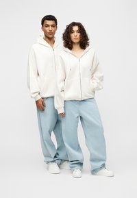 Λευκές φούτερ με φερμουάρ και φαρδιά εφαρμογή, σε συνδυασμό με ανοιχτόχρωμο μπλε παντελόνι με φαρδιά μπατζάκια και λευκά αθλητικά παπούτσια. Απλός σχεδιασμός, μαλακή υφή, χωρίς σχέδια.