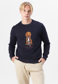 Felpa navy con una grafica testurizzata di un cane dorato e dettagli tartan. Presenta un collo a giro e polsini a costine. Logo "HACKETT" sotto.