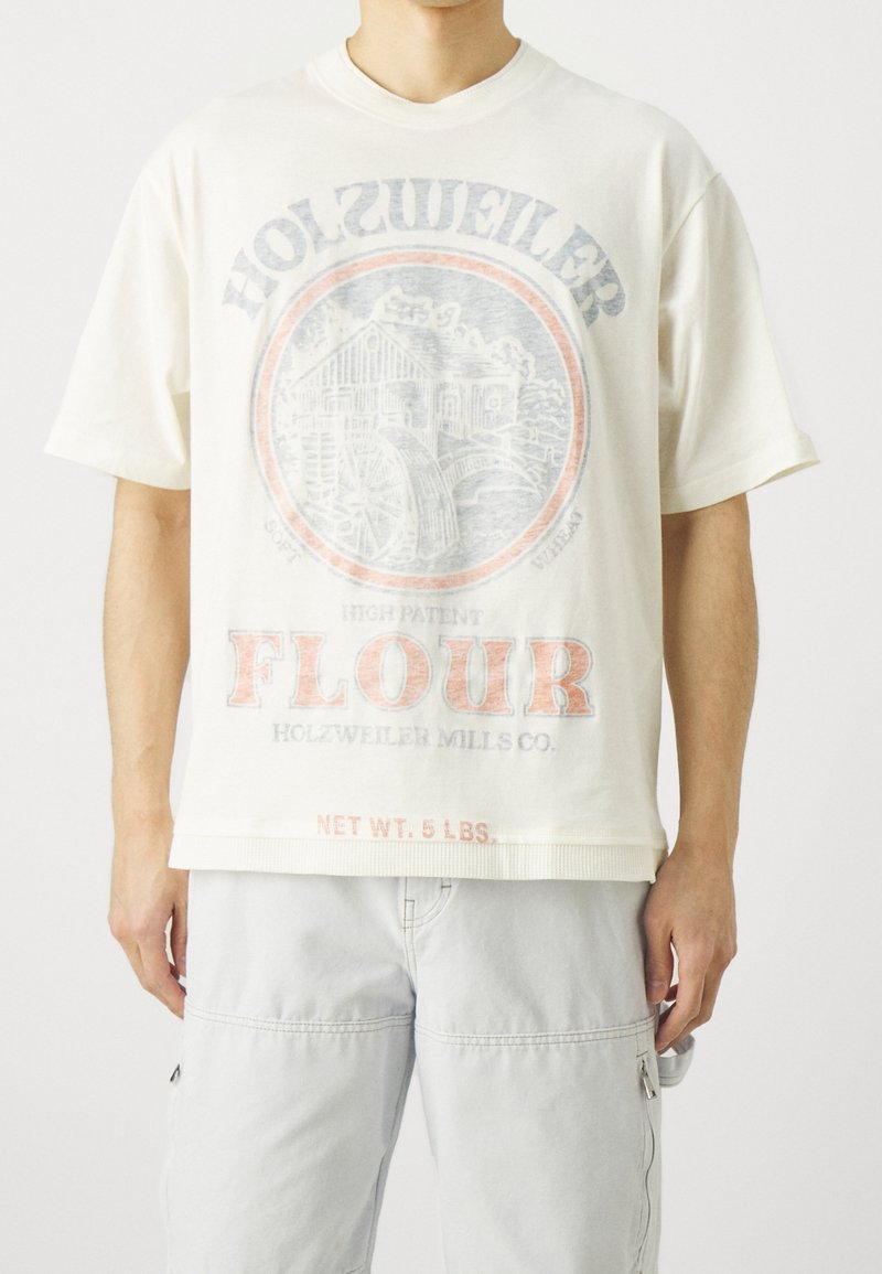 Personne portant un t-shirt blanc ample avec un logo de moulin à farine vintage délavé et le texte "HOLZWEILER FLOUR", ainsi que un short cargo de couleur claire.