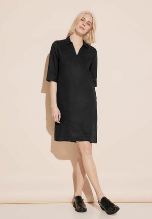 Schwarzes kurzärmeliges Kleid mit Kragen und lockerer Passform. Glatte Baumwolltextur, knielang, kombiniert mit schwarzen Schnürschuhen.