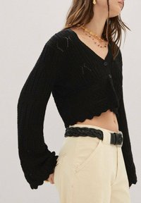 Cardigan noir court avec un motif de tricot texturé, en v et fermeture à boutons. Présente des manches longues avec des bords froncés, associé à un pantalon beige.