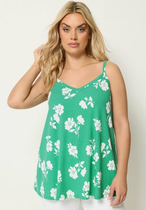 Lyshåret kvinde iført en grøn ærmeløs top med hvidt blomsterprint, poserer med den ene hånd, der rører ved hendes hår.