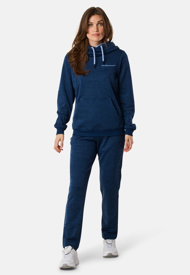 Kvindelig person står iført en marineblå hættetrøje og matchende sweats, samt hvide sneakers, mod en ensfarvet hvid baggrund.