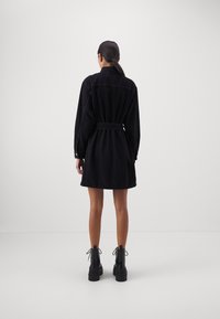 Vrouw gezien van achteren, gekleed in een zwart denimjurk met riem en lange mouwen, met zwarte veterlaarzen tot de enkel, staand tegen een effen achtergrond.