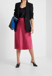 Svart blazer över en svart satinlinne, kombinerad med en rosa satinfotsidlång kjol och svarta klackade muletofflor; håller en blå och svart handväska.