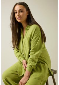 Camisa verde lima de botón con un bolsillo en el pecho y mangas largas, combinada con pantalones a juego. La tela parece texturizada y suave.
