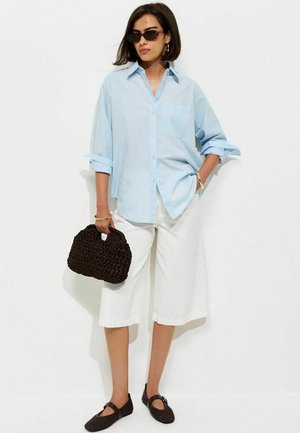 Vrouw met kort haar die een zonnebril draagt, lichtblauw oversized shirt, witte wijde shorts, houdt een zwarte gevlochten tas vast en draagt zwarte schoenen.