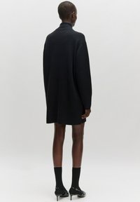 Schwarzes, übergroßes Strickkleid mit hohem Kragen, langen Ärmeln und lockerer Passform, hergestellt aus einem strukturierten Strickstoff. Abgerundet mit schwarzen Schuhen.