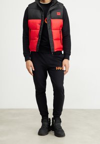 Homme portant une doudoune noire et rouge par-dessus un sweat-shirt noir et un pantalon de jogging avec un logo en flammes, assortis à des bottes de randonnée noires sur un fond uni.