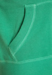 Nærbillede af grøn sweatshirt-stof med en kængurulomme med buet kant, synlig syning og ribbet struktur langs kanten.