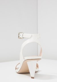 Bílé kožené sandály na vysokém podpatku s řemínkem kolem kotníku, se špičatou klínovou podrážkou a hladkou texturou. Spona na řemínku.