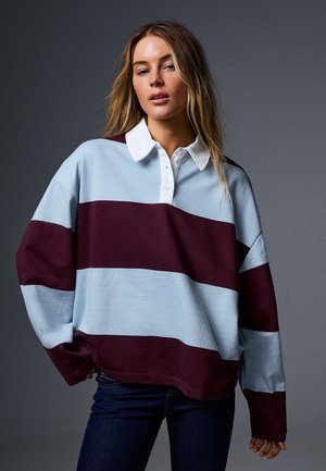 Femme aux cheveux lâches et ondulés portant un maillot de rugby rayé bleu clair et bordeaux avec un col blanc et un jean foncé sur un fond sombre.