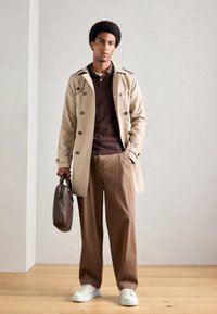 Uomo in piedi all'interno che indossa un trench beige, una polo marrone, pantaloni marroni, sneakers bianche e tiene una valigetta in pelle marrone scuro.