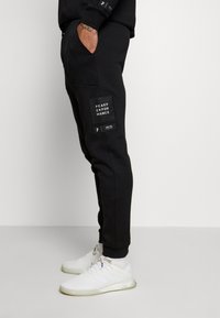Pantalon de survêtement noir à coupe décontractée, avec poches avant et un écusson en tissu portant le logo "PEAK PERFORMANCE". Le matériau semble doux et texturé.