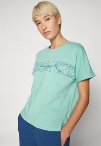Mintgrønn bomull T-skjorte med korte ermer, med et blått geometrisk Mizuno-logo-design over brystet. Vanlig passform.