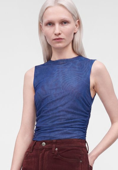 Jeune femme aux cheveux blonds platine et raides portant un haut bleu sans manches et un pantalon bordeaux sur un fond uni.