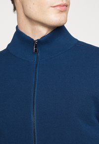 Marinblå zip-up tröja med hög krage, texturerad stickad tyg och en kontrasterande dragkedja. Närbild av halsringning och dragkedjedetaljer.