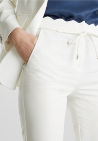 Personne portant un pantalon blanc avec une taille à cordon et un blazer blanc, main posée de manière décontractée dans la poche du pantalon, haut bleu marine visible.